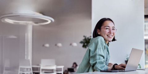 Eine lächelnde junge Frau sitzt vor einem Laptop an einem hellen und großräumigen Arbeitsplatz.